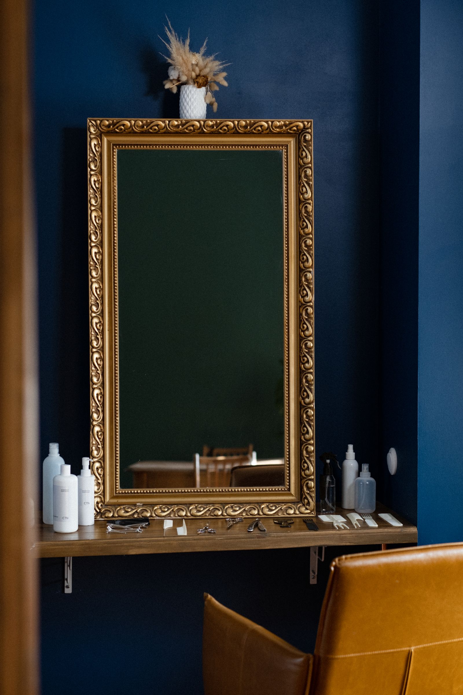 An ornate gold-framed mirror on a deep-blue wall above a wooden shelf holding plain white bottles, combs and scissors, with a tan leather chair edge below and a small dried-flower vase on top of the mirror